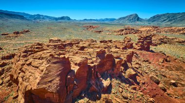 Geniş kızıl kaya kayalıkları, açık mavi gökyüzünün altında engebeli dağlarla çöl zemininden yükseliyor.