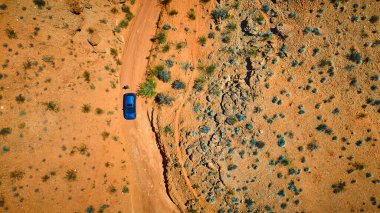 Mavi bir araç ve yalnız bir insan, parlak güneş ışığı altında engin bir kayalık çöl manzarasında virajlı toprak bir yolda dururlar.
