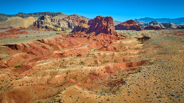 Canlı kırmızı kayalar, engebeli çöl arazisinin üzerinde, açık mavi gökyüzünün altında, dağınık kurak bitki örtüsüyle yükseliyor.