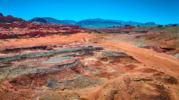 Uçsuz bucaksız kızıl kaya oluşumları engebeli çöl arazileri ve parlak mavi gökyüzünün altındaki uzak dağlar.
