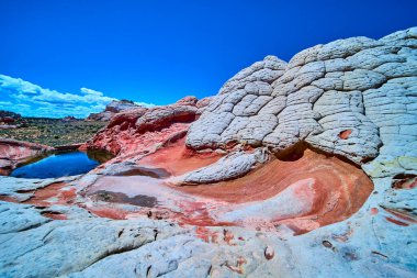 Arizona 'daki Vermilion Cliffs Ulusal Anıtı' nda parlak kırmızı ve beyaz kumtaşı oluşumları, çarpıcı çizgiler ve çölde canlı mavi bir gökyüzünün altında sakin bir havuz bulunmaktadır.