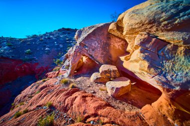 Canlı kırmızı kumtaşı katmanları ve engebeli kaya oluşumları, Nevada 'daki Gold Butte Ulusal Anıtı' nda açık mavi gökyüzüne karşı durmakta ve seyrek çöl bitkileri dramatik sahneyi vurguluyor.