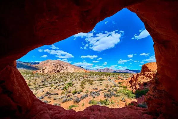Kızıl kaya kemeri oluşumu, Nevada 'nın engin çöl ve engebeli tepelerini, canlı mavi gökyüzü ve dağınık bulutlarla çevreleyerek dramatik bir ölçek ve macera hissi yaratır.
