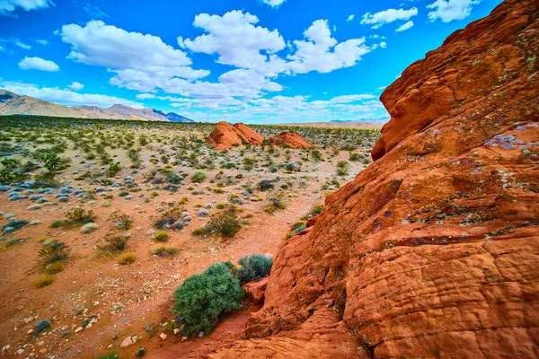 Canlı kırmızı kumtaşı kayaları, yerli bitkiler ve Nevada Ulusal Anıtı 'ndaki uzak dağlarla dolu bir çöl arazisinin üzerinde yükseliyor. Doğal bir merak ve açık alan hissi yaratıyor.