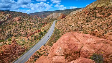 Kanyon Noktası ve Catstair Canyon Utah 'taki canlı kırmızı kaya oluşumlarından geçen dolambaçlı bir otoyolun hava görüntüsü açık yolda tek bir araba açık mavi gökyüzü ve dağınık bulutlar altında