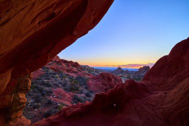 Nevada 'daki Gold Butte Ulusal Anıtı' nda canlı kırmızı kumtaşı oluşumu, uzak dağlar ve Whitney Pocket yakınlarında renkli günbatımı gökyüzü ile bir çöl sahnesini çerçeveler.