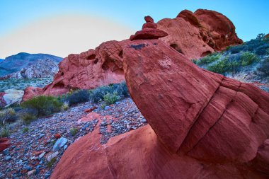 Göz alıcı bir cairn, Nevada 'daki Gold Butte Ulusal Anıtı' ndaki canlı kırmızı kayaların üzerinde çöl bitkileri ve engin gökyüzü altında dramatik taş oluşumlarıyla çevrili duruyor.