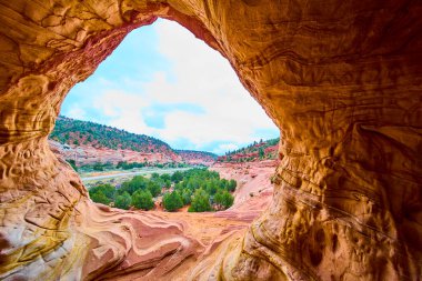 Kanab Utah yakınlarındaki dokulu bir kumtaşı mağarasının içinden bakıldığında kırmızı kaya tepeleri, canlı ardıç ağaçları ve parlak güneş ışığı altındaki uzak mesafelere sahip dramatik bir çöl manzarasını gözler önüne seriyor.
