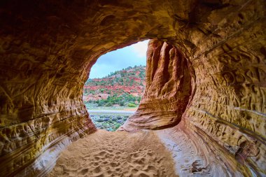 Kanab Utah yakınlarındaki dokulu bir kumtaşı mağarasının içinden bakıldığında kumlu zeminde ayak izleri ve Amerika 'nın güneybatısında kırmızı kayalar ve yeşilliklerle aydınlatılmış çarpıcı bir çöl manzarası görülüyor.
