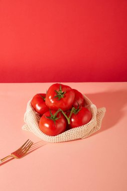 Sulu kırmızı domatesler, koyu pembe arka planı olan pastel pembe bir masanın üzerindeki metal çatalın yanında, kumaş bir sepetin içinde saklanır. Reklam için boşluk kopyala.
