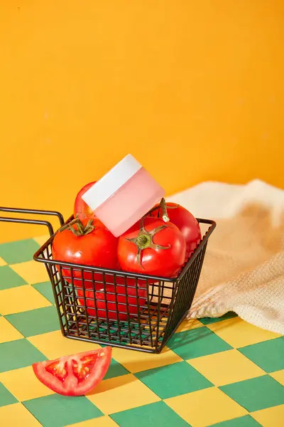 An unlabeled cosmetic jar is placed on top of juicy red tomatoes in a wire mesh basket. Unique background with turquoise and yellow checkered pattern. Concept of vegan cosmetics.