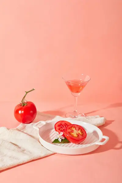 Sliced tomatoes and a camellia flower are placed on a round ceramic tray, next to a tomato and a cocktail glass containing tomato juice on a pink background. Copy space for ads.