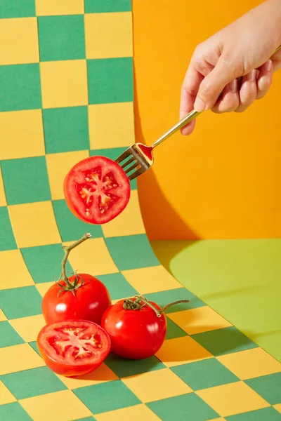 手はトマトの半分に固定された金属製のフォークを保持しています ジューシーな赤いトマトは 緑と黄色の2つの主要な色でチェッカーされた背景に表示されます 新鮮な野菜とアートスペース — ストック写真