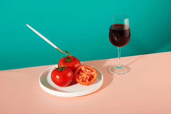 金属製のフォークは 白いセラミックプレートに新鮮なトマトに詰め込まれています その隣には赤ワインが置かれています ターコイズとパステルピンクの背景 新鮮な野菜を使った広告シーン — ストック写真