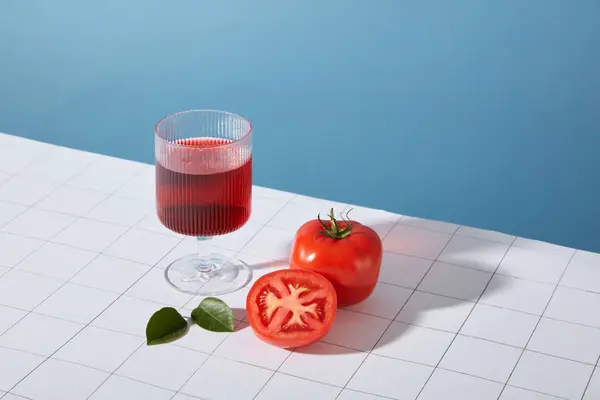 透明な赤い液体を含むガラスは 青と白の背景に新鮮なトマトと緑の葉の隣に配置されています トマトはB1 B9などの多くのBビタミンが含まれています — ストック写真