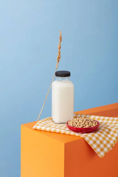 Blank label bottle of milk and a ceramic dish of soybeans are displayed on a white and orange checkered fabric. Nut milk is rich in vitamins and minerals