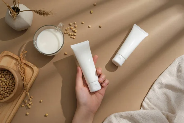 Female hand model is holding an empty label tube. A cup of milk and a bowl of soybeans displayed. Soybean (Glycine max) is very good for skin and health