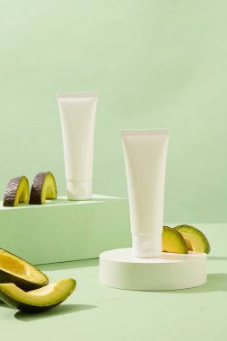 Podyumda etiketsiz beyaz plastik tüplerin ön görüntüsü, yeşil arka planda avokado dilimleriyle süslenmiş. Avokado 'daki kozmetik ürünleri reklamı sahnesi