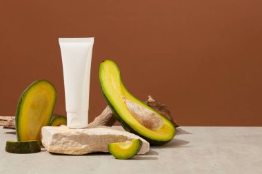 Kahverengi arka planda, taze avokado ve kuru dal parçalarıyla süslenmiş, taş kürsüye yerleştirilmiş beyaz plastik tüpler. Ön görünüm, dizayn ve kopyalama alanı için boşluk