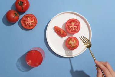 Beyaz bir tabakta bir bardak domates suyuyla süslenmiş birkaç dilim domates vardı. El modeli çatal tutuyor. Domatesler iltihap önleyici bileşikler bakımından zengindir.