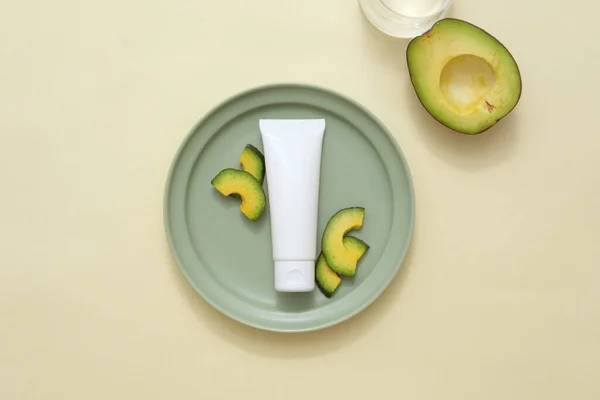 Top view of white cosmetic tube placed on round green dish with fresh avocado slices on beige background. Plastic tube mockup for cosmetic, without label for design packaging.