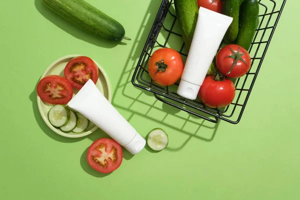 Two blank label tubes in white color displayed with a dish and a basket of tomatoes and cucumbers. Mockup of skin care cosmetic tube of beauty facial