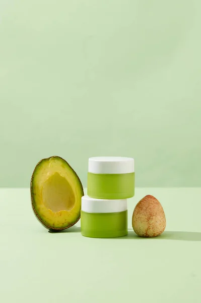 Front view of two green cosmetic cream jars displayed on pastel background with halves of avocado and seed. Minimalist scene for advertising and branding cosmetic with blank space for design