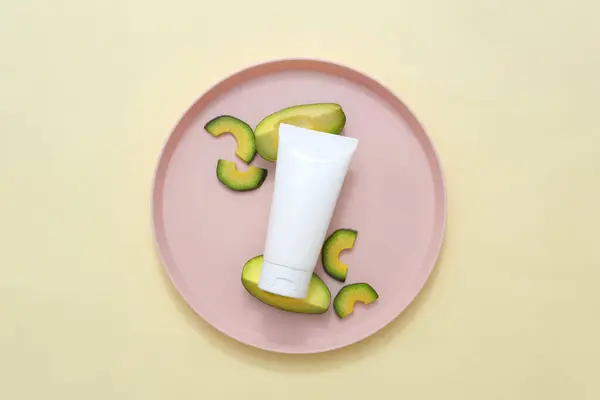 Beauty product packaging design templates based on avocado. On a beige background, a white plastic tube decorated with fresh avocado slices on pink ceramic dish. Top view, advertising photo