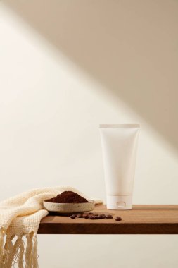 Tahta kaidede beyaz plastik bir tüp, kahve çekirdekleri, seramik bir kase kahve tozu ve beyaz arka planda bej kumaş sergileniyor. Kahve telvelerinin güçlü iltihap önleyici özellikleri vardır.