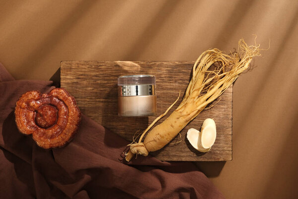 On a wooden podium, a cosmetic jar decorated with root and slices of ginseng, lingzhi mushroom and brown cloth on brown background. Concept for cosmetics extracted from rare herbs. Top view