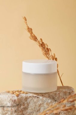 Bej arka planda taş bir podyumda etiketsiz bir kozmetik kavanozu ve pirinç sergileniyor. Pirinç kepeği özü doğal bir özüttür ve cilt bakımında birçok kullanım alanı vardır..