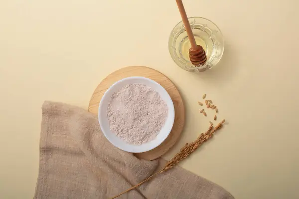Rice bran powder is displayed on a ceramic plate on a wooden platform. A honey drizzle is placed in a glass of water. Rice bran powder is very effective in tightening pores and removing dirt.
