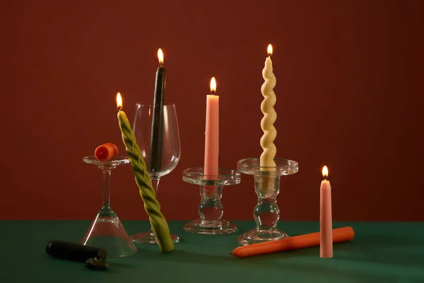 Burning colored candles are placed on glass candlesticks on a deep green and brown background. Candles are used for lighting and are an indispensable item on birthdays.