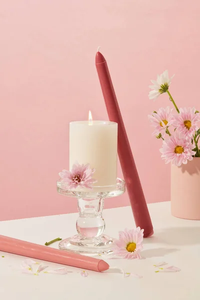 燃烧的蜡烛的前视图放在玻璃烛台上 周围装饰着彩色的蜡烛和淡淡的花朵 背景是白色和粉色的 — 图库照片