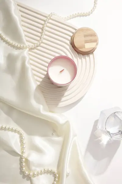A string of pearls, a jar of scented candles, a silk cloth, a glass of water and a dome-shaped podium on a white background. Sophisticated space with candles.