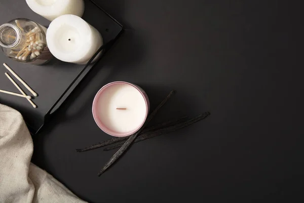Two white candles and a bottle of cotton swabs are displayed on a black tray. A jar of scented candles is placed on the table with a black background. Candle advertisement with minimalist background.