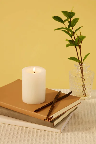 一支点燃的蜡烛放在笔记本上 旁边是一个玻璃花瓶 绿色的叶子在桌子上 背景是黄色的 大多数蜡烛都是石蜡做的 — 图库照片