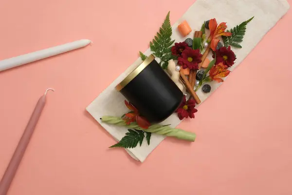 Unbranded black scented candles beside fresh flowers, colored candles, green leaves and cinnamon on a pink background with a white cloth. Ideal mockup for scented candle advertising.