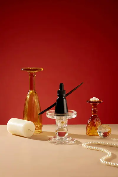 一个黑色的血清瓶子放在玻璃烛台上 装饰玻璃杯 白色的蜡烛和一串珍珠陈列在米色的桌子上 红色背景 复制的创造性空间 — 图库照片