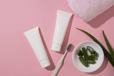 Dilimlenmiş aloe vera seramik bir tabakta sergilenir. Etiketlenmemiş iki kozmetik tüp pembe arka planda bir havlu ve diş fırçasıyla yerleştiriliyor. Vejetaryen kozmetik reklamları.
