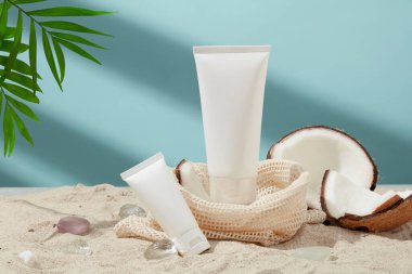 Beyaz kumlu simüle edilmiş bir plaj arka planında, iki tüp beyaz losyon, taze hindistan cevizi, yeşil yapraklar ve bir kumaş sepet sergilenir. Vejetaryen kozmetik reklamı sahnesi.