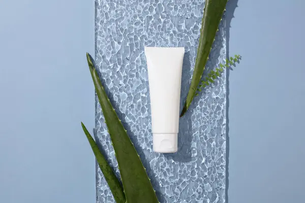 Close-up of an unlabeled cosmetic tube displayed on a water wave-effect glass platform with aloe vera leaves. Blue background. Fresh space for advertising natural cosmetics.