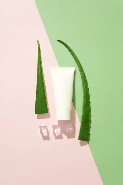 On a green-pink color scheme, a white cream tube is placed in the middle, next to fresh aloe vera and ice. Copy space for advertising cosmetics with natural ingredients.
