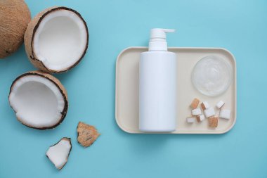 Pastel mavi arka plan, seramik tabak ve taze hindistan cevizi. Reklam için merkeze etiketsiz bir beyaz duş jeli şişesi yerleştiriliyor. Tropikal meyveli kozmetik ürünler.