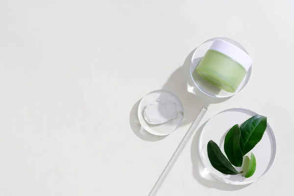 A jar of eye cream is placed on a glass platform, a few green tea leaves are decorated next to it on a white background. Green tea essence helps reduce eye puffiness and eliminate dark circles.