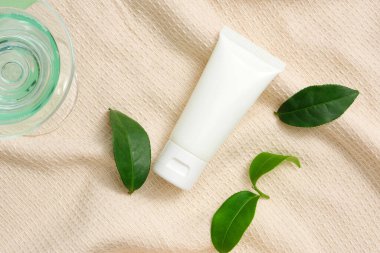 Bir bardak şeffaf yeşil sıvı kozmetik tüp ve pastel kumaş zemin üzerinde yeşil çay yaprakları ile sergilenir. Reklam ve markalaşma için mükemmel bir model..