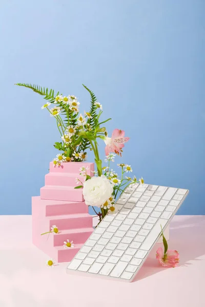 Front view of a white computer keyboard is placed next to pink stairs and assorted flowers on a blue-pink background. Scene for advertising and magazine posting.
