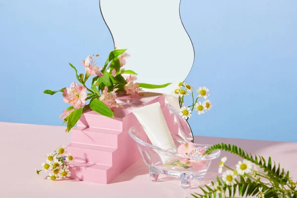 The front of a white unlabeled cosmetic tube is placed in a mini glass tub, next to it are fresh flowers and some other props on a pastel background. Advertising natural cosmetics.