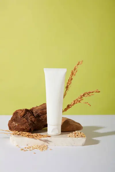 Front view of an unlabeled cosmetic tube is placed on a white platform with whole grain rice and a stone slab. Vegan cosmetic concept with main ingredient from rice bran.