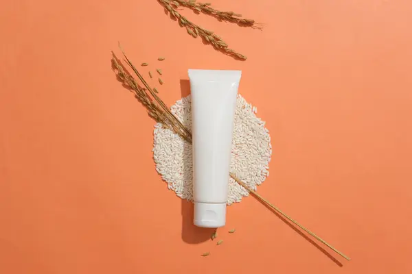 A tube without label placed on a pile of rice and decorated with wheat ears. Orange background. Empty label for cosmetic product mockup